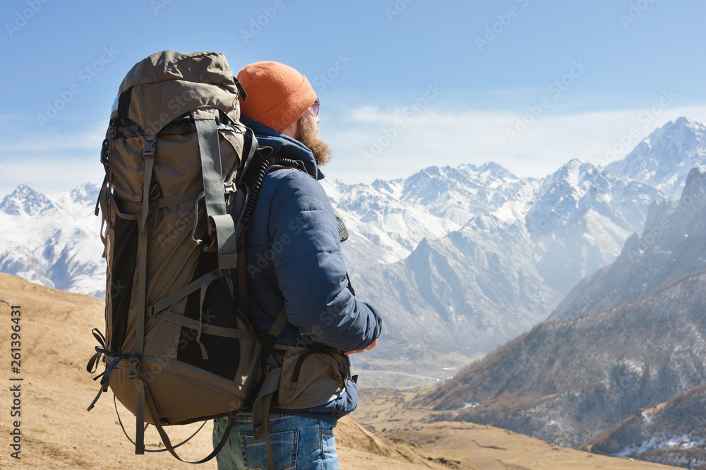 Portrait of a bearded man in sunglasses and a warm jacket with a backpack on his back against the background of snowy mountains on a sunny day. The concept of tourism and travel