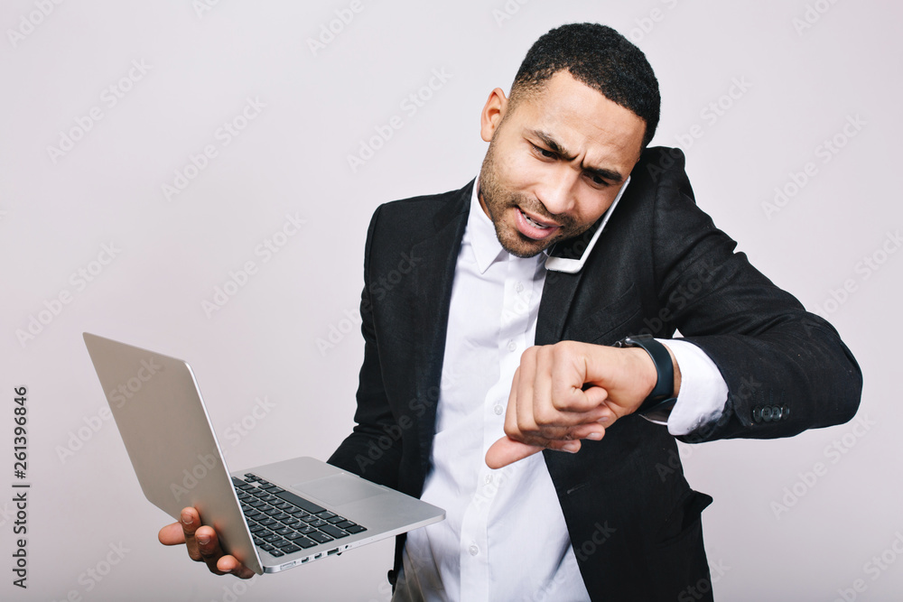 Portrait hard-working busy young man in white shirt, black jacket talking on phone and looking at watch on white background. Stylish businessman, working witj laptop, time for work, meeting