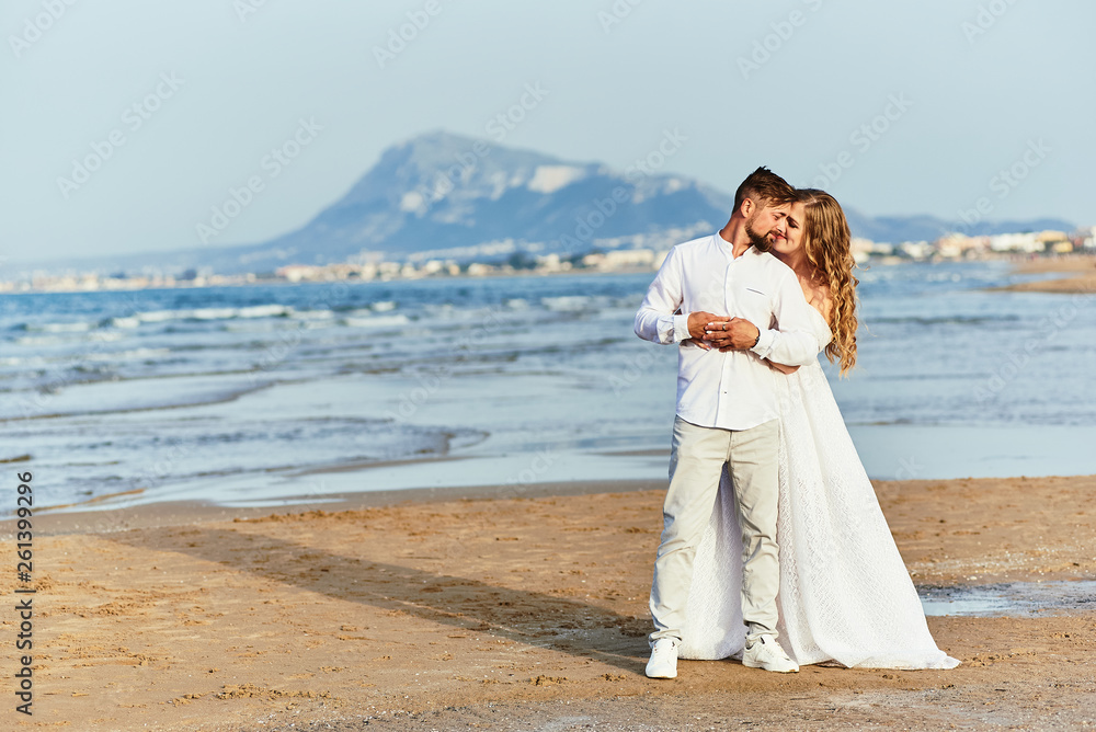 Young blonde bride in white wedding dress embraces groom on the beach Young blonde bride in white wedding dress embraces groom on the beach
