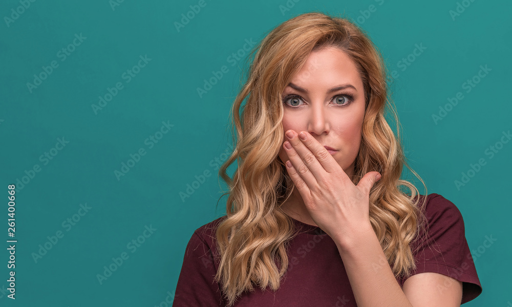 Attractive blonde holds hand at mouth, surprised or closed, on a blue background. Attractive blonde holds hand at mouth, surprised or closed, on a blue background.