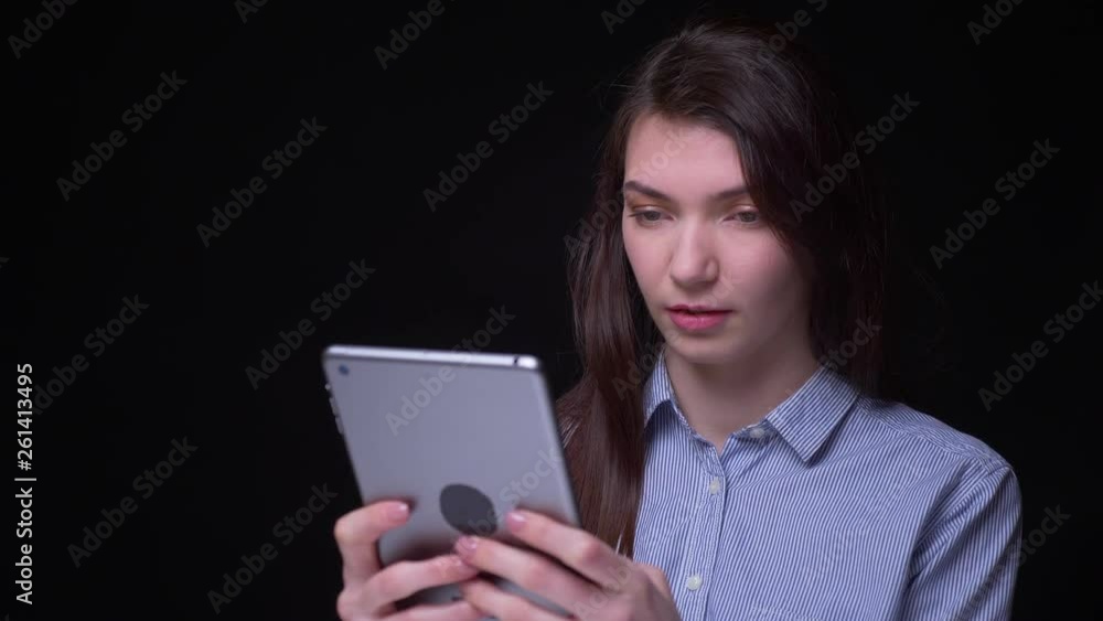 Portrait of young brunette businesswoman in blue blouse smilingly watching into tablet at black background. Portrait of young brunette businesswoman in blue blouse smilingly watching into tablet at black background.