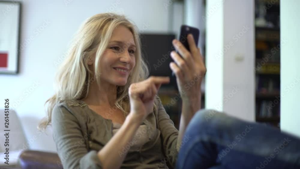 Medium shot of a mature woman taking selfie and texting on her smart phone while sitting on the chair at home Medium shot of a mature woman taking selfie and texting on her smart phone while sitting on the chair at home