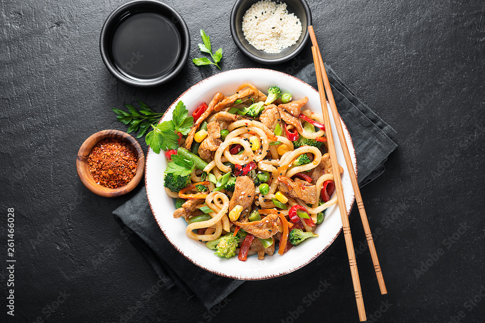 Udon stir fry noodles with pork meat and vegetables in a white plate on black stone background.
