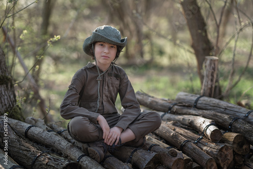 A boy in a coveralls and a hat sitting on a pile of sawn logs in the forest.