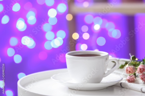Cup of hot drink and flowers on table against blurred lights, space for text