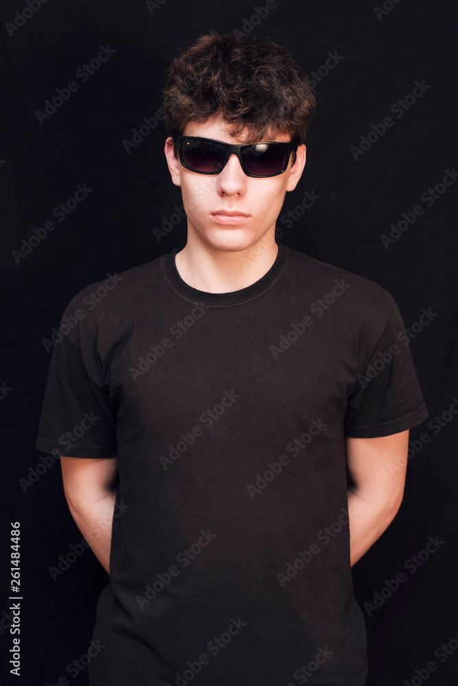 Fototapeta premium A boy of an adolescent age in a black shirt and in glasses from the sun on a black background. The boy has a serious face