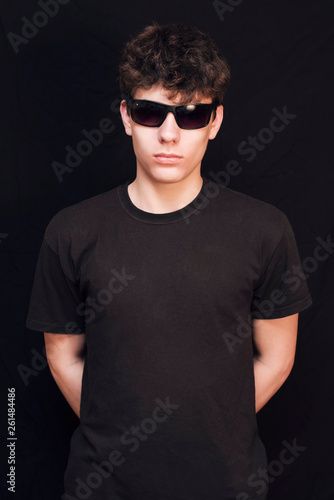 A boy of an adolescent age in a black shirt and in glasses from the sun on a black background. The boy has a serious face