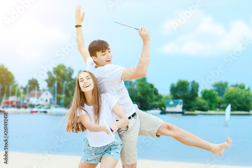 Young smiling man and woman in light clothes are playing in matkot on nature. Couple in love is having fun on beautiful beach with yachts and sailing boats on background. Summer activities concept.