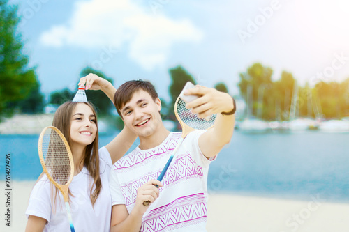 Young couple in love female girl and man male teenagers adults playing in badminton with rackets and shuttlecock near water, river, lake at hot summer day at beach and doing funny selfie by smartphone © Monstar Studio