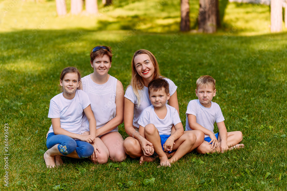 Two happy young mothers and three children in white t-shirts sit on a green lawn in the summer. Large Caucasian family on vacation Two happy young mothers and three children in white t-shirts sit on a green lawn in the summer. Large Caucasian family on vacation