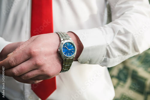 A man in a white shirt and red tie puts a wristwatch on his arm.