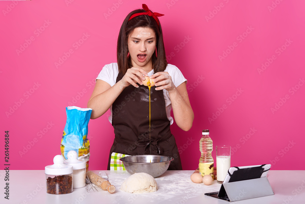 Close up portrait of woman smashes and punches egg for cake preparation. Concept of baking and preparing for Easter holiday. Adds eggs as ingredient to dough. Baking hot cross bun over pink background