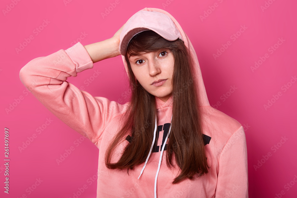 Image of brunette girl looks at camera with serious and thoufgtfull facial expression, wearing casual pink hoody and cap isolated on rose background, teenager poses in photo studio. Modeling concept. Image of brunette girl looks at camera with serious and thoufgtfull facial expression, wearing casual pink hoody and cap isolated on rose background, teenager poses in photo studio. Modeling concept.