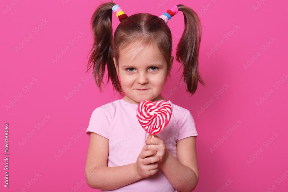 Beautiful fair haired child stands straight, looks directly at camera, holds colourful lollipop in hands, having frowny face, has no desire to share candy with anybody. Copy space for advertisement.