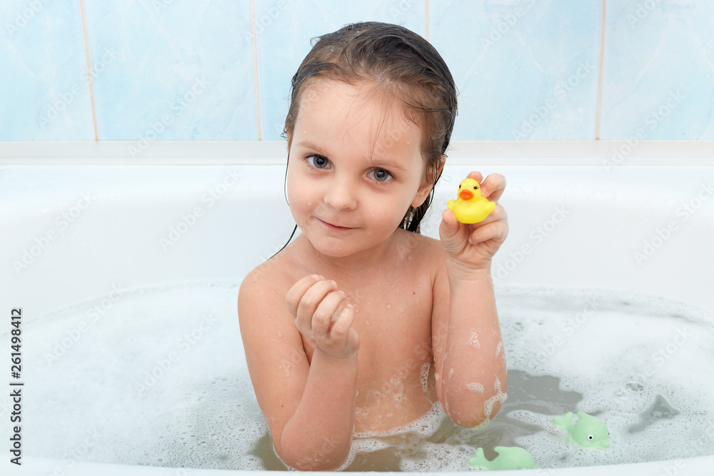 Close up portrait of happy charming little girl sitting in bath tub plays with yellow duck in bathroom. Baby bathing with toys in bath full of foam, showing toy to mother, looks seriously at camera Close up portrait of happy charming little girl sitting in bath tub plays with yellow duck in bathroom. Baby bathing with toys in bath full of foam, showing toy to mother, looks seriously at camera