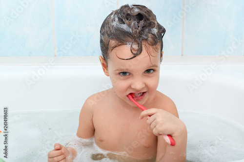 Portrait of small charming baby girl brushing her teeth  sitting in bathtub  caring about teeth health. Magnetic little girl washes over light blue bathroom wall. Hygiene and children concept.