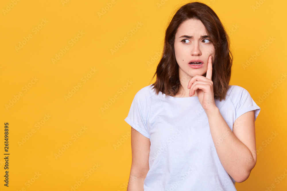 Portrait of beautiful brunette pensive young woman in light clothes looking aside, keeps fore finger on cheek, isolated on yellow wall background in studio. People sincere emotions, lifestyle concept. Portrait of beautiful brunette pensive young woman in light clothes looking aside, keeps fore finger on cheek, isolated on yellow wall background in studio. People sincere emotions, lifestyle concept.