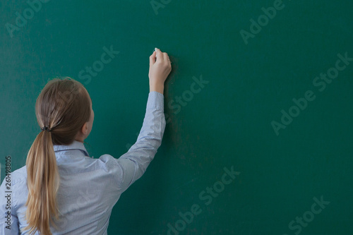 Rear view of a student or teacher with long brunette hair writing on a blank green blackboard or chalkboard with copyspace