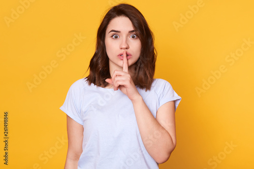 Slim attractive young lady stands straight isolated over bright yellow background, holding forefinger finger on her lips, making hint of being silent, opens her eyes wide, wears white casual t shirt.
