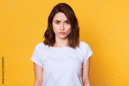 Portrait of attractive miserable girl having disappointed look on her face, looking directly at camera. Emotional charming model poses isolated over yellow background. People and emotions concept.