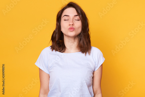 Young magnetic brunette lady standing with closed eyes, protruding her lips, making kissing sign, looks romantic. Slender energetic model has peaceful facial expression. Emotional state concept.