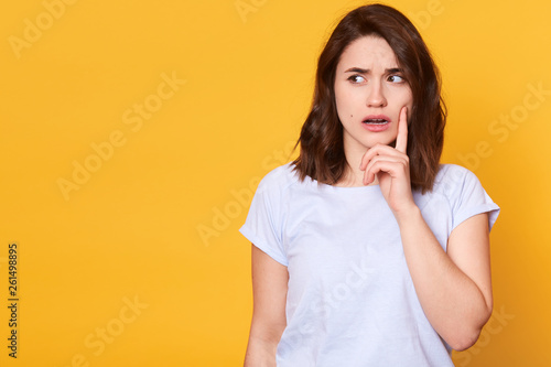 Portrait of beautiful brunette pensive young woman in light clothes looking aside, keeps fore finger on cheek, isolated on yellow wall background in studio. People sincere emotions, lifestyle concept.