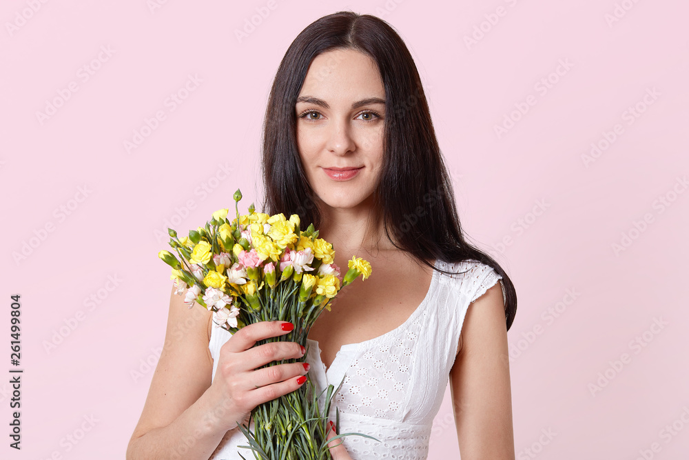Charming beautiful woman holds yellow pink flowers with one hand, looking directly at camera, feels pleased. Smiling brunette model poses in white summer dress. Copy space for advertisement.