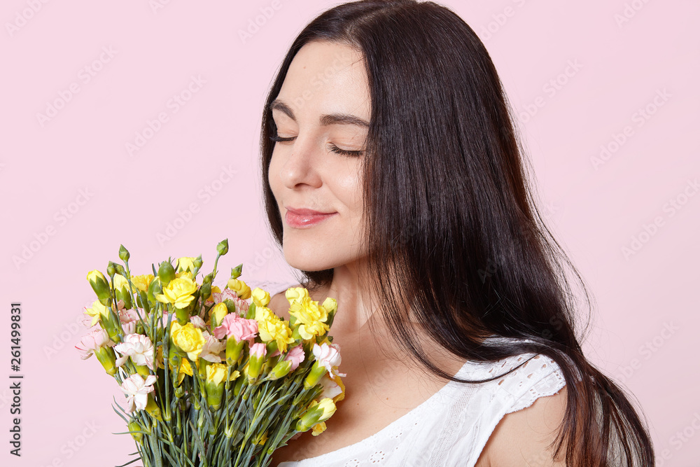 Close up portrait of black haired adorable young lady standing with closed eyes, holding early spring flowers, enjoys fantastic smell, looks delighted and peaceful. People and emotions concept.