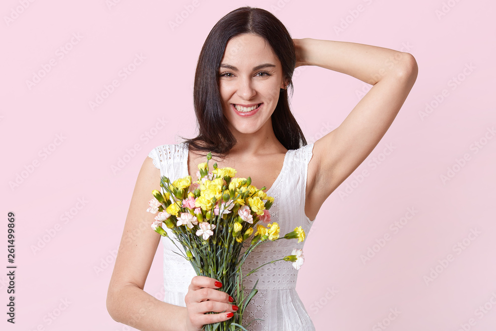 Portrait of a happy cheerful young woman in summer white dress holding colourful flowers in one hand, fixing her hair, looking directly at camera. Smiling model poses isolated over pink background. Portrait of a happy cheerful young woman in summer white dress holding colourful flowers in one hand, fixing her hair, looking directly at camera. Smiling model poses isolated over pink background.