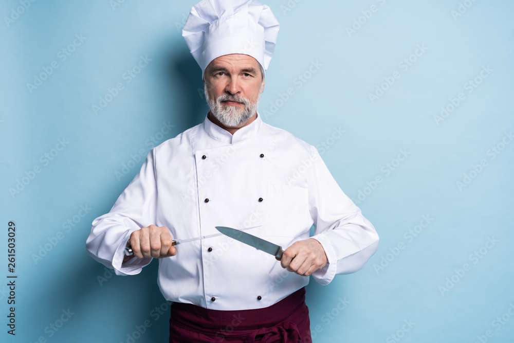Portrait of a mature chef cook holding knifes isolated on a blue background. Portrait of a mature chef cook holding knifes isolated on a blue background.