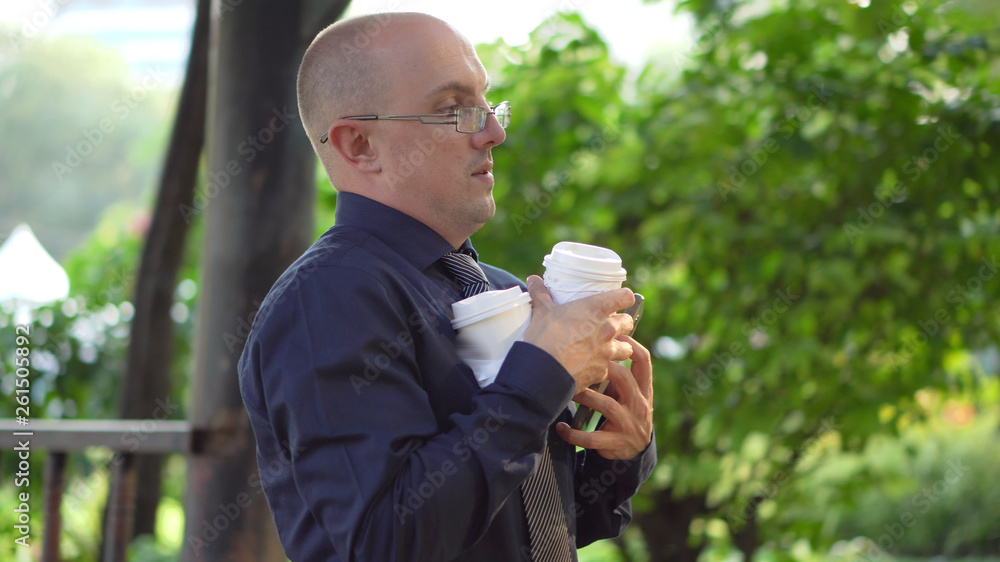 Overworked Young Caucasian Business Man With Two Cups of Coffee Taking Work Phone Call On Lunch Break in Park Overworked Young Caucasian Business Man With Two Cups of Coffee Taking Work Phone Call On Lunch Break in Park