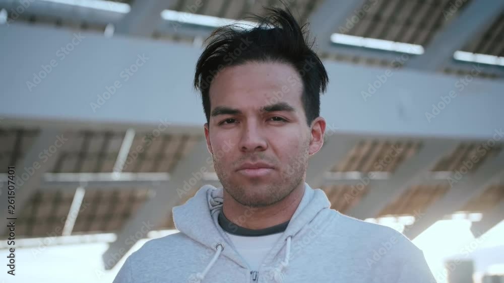 Portrait of young Hispanic man with a serious pensive look at the camera, standing in the street at the sunset, concrete construction on the background, dressed in grey hoodie Portrait of young Hispanic man with a serious pensive look at the camera, standing in the street at the sunset, concrete construction on the background, dressed in grey hoodie