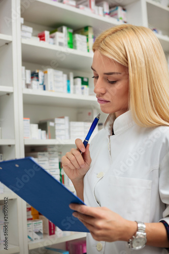 Serious young female pharmacist with blond hair holding a clipboard and reading. Shelves full of medications in the background
