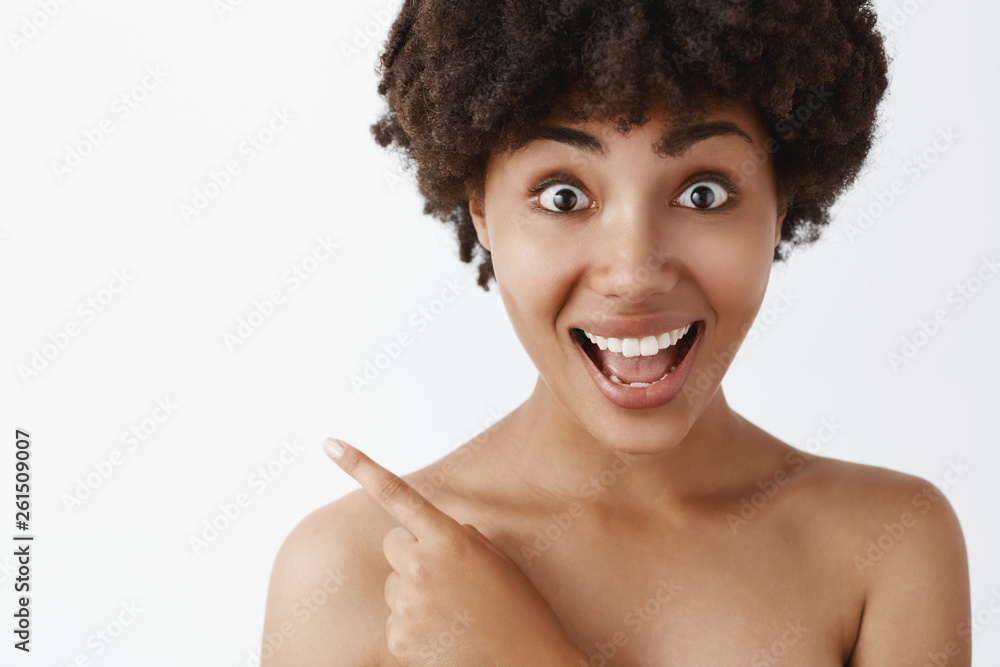 Amused and thrilled african american female model with curly haircut standing naked with clean skin, pointing at upper left corner, smiling joyfully, being impressed and satisfied with product Amused and thrilled african american female model with curly haircut standing naked with clean skin, pointing at upper left corner, smiling joyfully, being impressed and satisfied with product