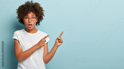 Indoor shot of impressed surprised beautiful woman with curly hair, dark skin, points both fore fingers at upper right corner, isolated over blue studio wall, shocked by high price on item in shop