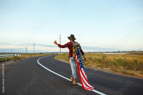 4th of July. Fourth of July. American with the national flag. American Flag. Independence Day. Patriotic holiday. The man is wearing a hat, a backpack, a shirt and jeans. Beautiful sunset light. 