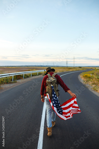 Fototapeta Naklejka Na Ścianę i Meble -  4th of July. Fourth of July. American with the national flag. American Flag. Independence Day. Patriotic holiday. The man is wearing a hat, a backpack, a shirt and jeans. Beautiful sunset light. 