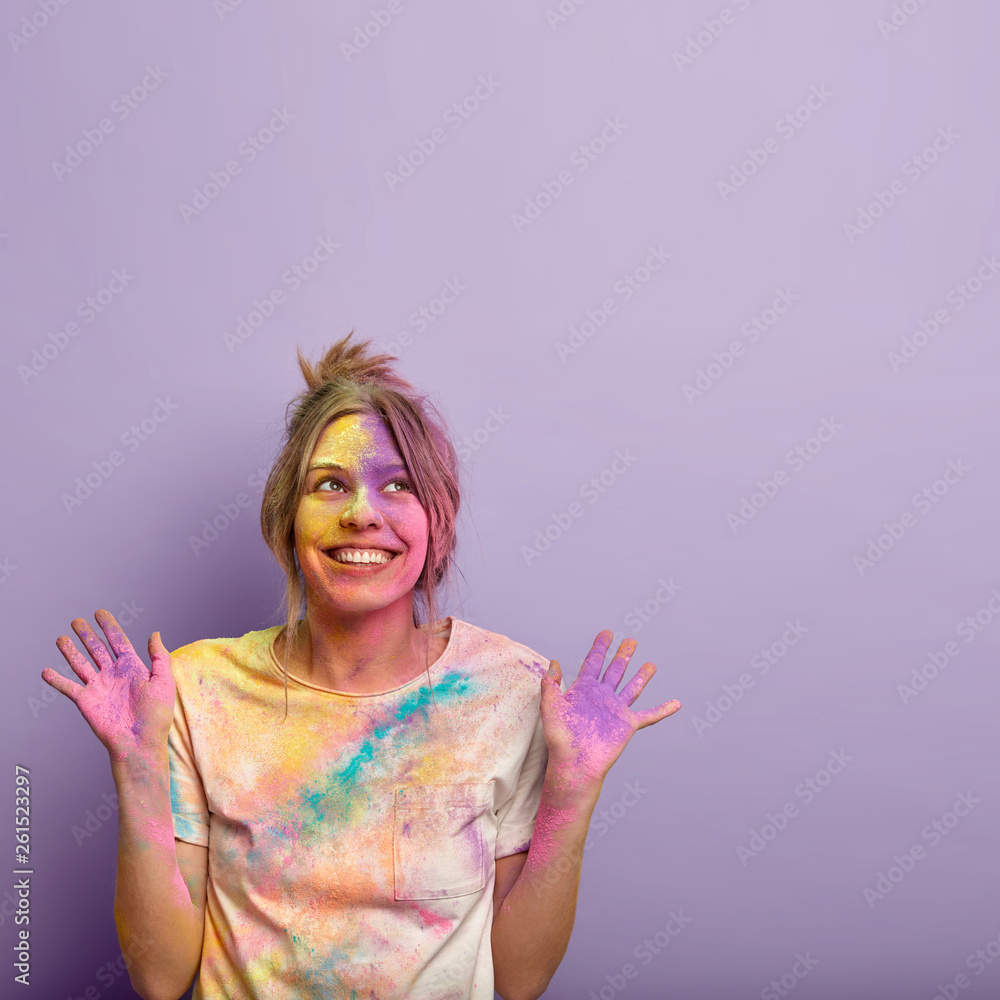 Isolated shot of glad dreamy young woman focused upwards, raises both hands and shows colored palms, smiles positively, celebrates Holi Festival of Colours, free space above for your information