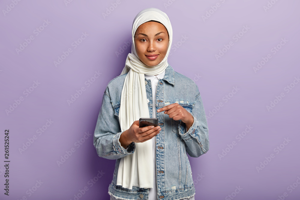Φωτογραφία Isolated shot of pleased mixed race young woman with dark skin, follows Muslim religion, points at mobile phone screen, asks to read internet news on website, isolated over purple background