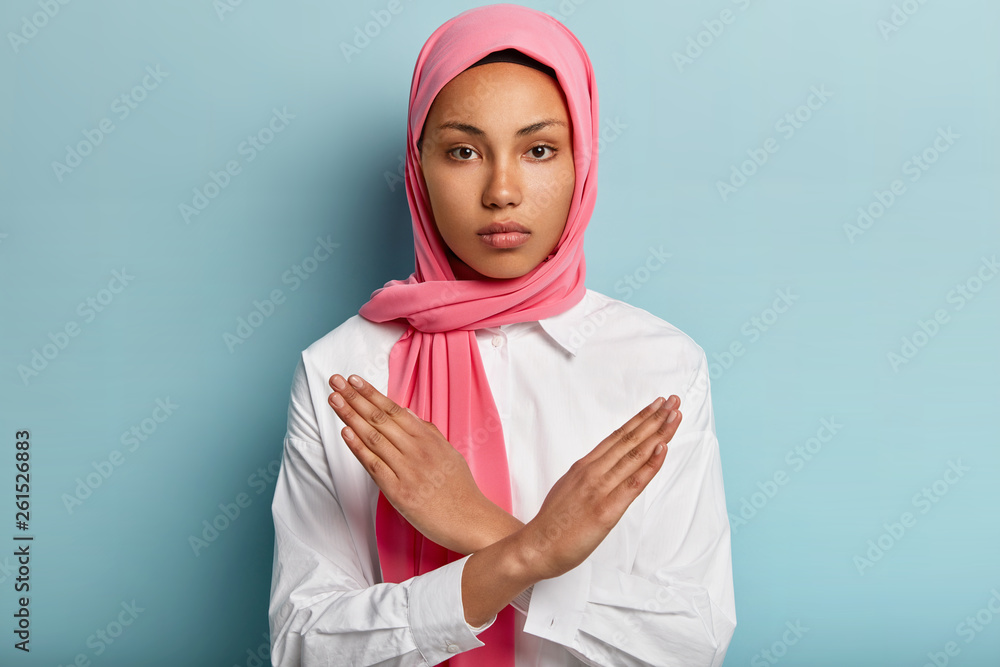 Indoor shot of serious Muslim woman makes denial hand gesture, keeps arms crossed over chest, demonstrates stop sign, wears headscarf, follows religious dress code, isolated over blue background Indoor shot of serious Muslim woman makes denial hand gesture, keeps arms crossed over chest, demonstrates stop sign, wears headscarf, follows religious dress code, isolated over blue background