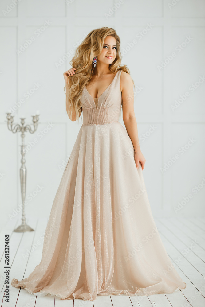 Beautiful blonde woman with greek hairstyle in beige powdery atlas wedding dress posing in studio room. Beautiful blonde woman with greek hairstyle in beige powdery atlas wedding dress posing in studio room.
