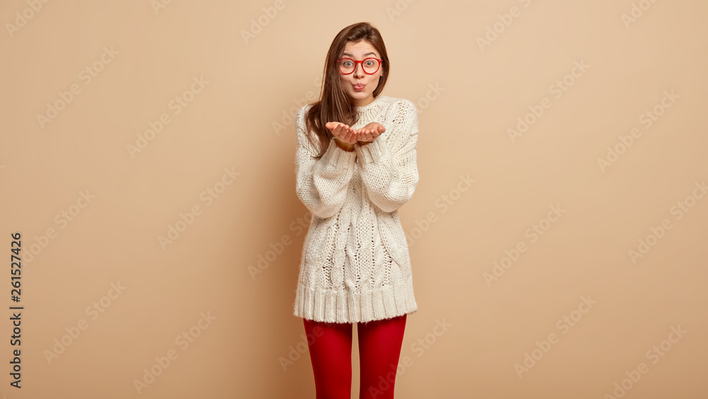 Φωτογραφία Adorable young European female model shows air kiss, protrudes palms at camera, expresses love to someone on distance, dressed in casual clothes, models over brown background
