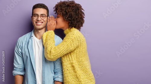 Afro American female whispers secret to boyfriend who has cheerful expression, gossip together, wear casual clothes, stands against purple background with blank space. Diverse couple indoor. photo