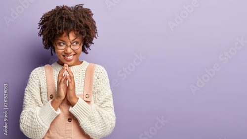 Foto Smiling dark skinned woman steeples fingers has great plan or intention, wears jumper and overalls, looks curiously away, poses against purple studio wall with free space
