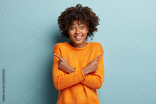 Pick one thing. Optimistic dark skinned female with glad expression  says both choice is great  crosses hands over chest  points at different sides  wears orange casual jumper  isolated on blue wall