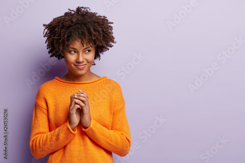 Pleased Afro American woman makes up plan  has intriguing look aside on free space  keeps palms together  looks away  wears orange jumper  isolated over purple background. People  intention concept.