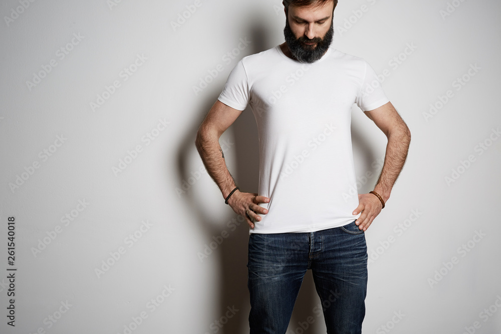 Young brutal tattooed bearded guy poses in blue jeans and blank white t-shirt premium summer cotton, on white background Young brutal tattooed bearded guy poses in blue jeans and blank white t-shirt premium summer cotton, on white background