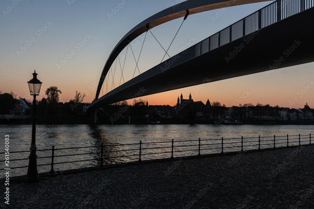 Obraz premium Sunset in downtown Maastricht seen from the riverside with a view on the historic skyline with the churches and hoge brug (high bridge)