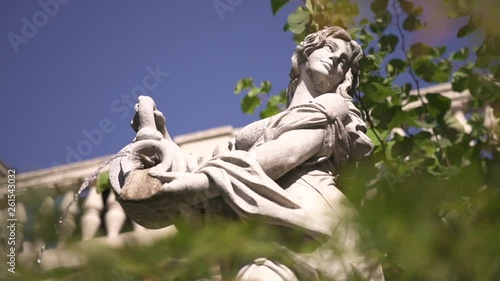 Classical gypsum figure holds amphora with water that drops on grass in front of baluster terrace in a green garden on clear blue sky background. Sculpture garden with trees, fir-trees and bushes.