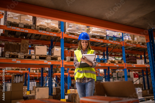 Warehouse female worker checking inventory in distribution warehouse. Smiling woman holding checklist.